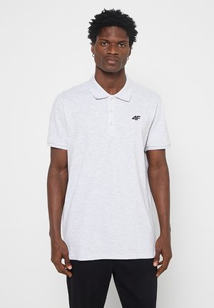 Man met kort krullend haar, die een lichtgrijs poloshirt met een klein zwart "4F"-logo draagt, staand tegen een effen witte achtergrond.