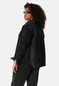 Zwarte, zachte stoffen shirt met een losse pasvorm en wijde mouwen. Heeft een kraag en knopen, gedragen over bijpassende zwarte broek.
