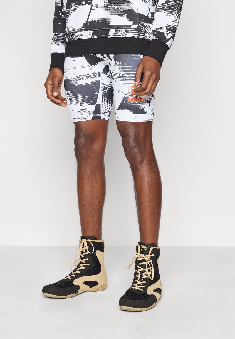 Ασπρόμαυροι σορτς με σχέδιο και πορτοκαλί λογότυπο, συνδυασμένοι με αθλητικά παπούτσια υψηλού τύπου σε μαύρο και μπεζ χρώμα με σχέδιο από δίχτυ.