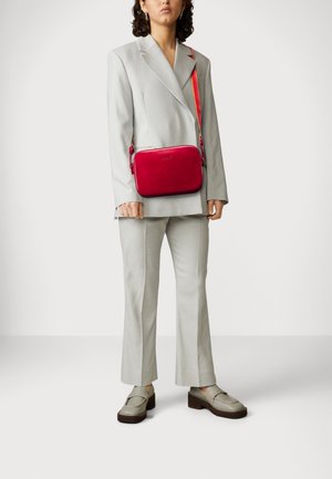 Röd läder crossbody-väska med rektangulär form, dragkedjestängning och märkeslogotyp framtill. Bärs med en ljusgrå kostym och beige skor.
