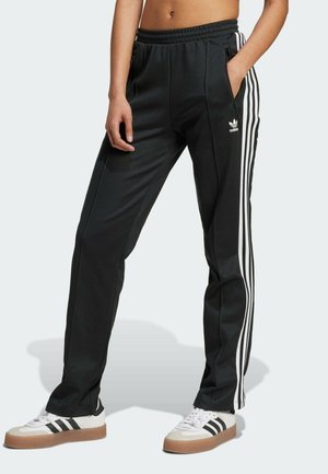 Personne portant un pantalon de survêtement Adidas noir avec des bandes blanches et des baskets blanches Adidas avec des semelles en gomme, debout sur un fond clair.