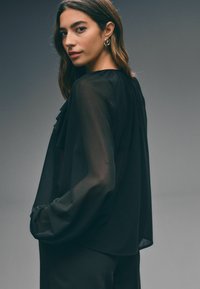 Sort, gennemsigtig bluse med lange ærmer og flæsedetalje foran, der har en løs pasform og afslappede ærmer. Glat tekstur.