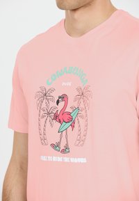 Vaaleanpunainen puuvillapaita, jossa on kuva auringonpaisteessa olevan flamingon, joka käyttää aurinkolaseja ja pitää surflautaansa, ympärillään palmuja ja leikkisiä fontteja.