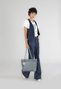 Chaleco de denim y jeans acampanados en azul oscuro, combinados con una camiseta gráfica blanca. Sosteniendo una bolsa de piel gris claro con herrajes dorados.