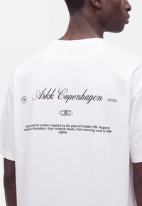 Άτομο που φοράει λευκό t-shirt Arkk Copenhagen με μαύρο κείμενο και λογότυπο στην πλάτη, εμφανιζόμενο από πίσω και πλαϊνή προφίλ.
