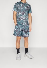 Hombre vestido con pantalones cortos y camisa de manga corta de estampado abstracto en verde, negro y blanco a juego, con calcetines negros y zapatillas blancas, de pie con las manos en los bolsillos.