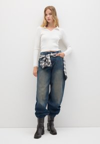 Giovane donna in top bianco a maniche lunghe, jeans blu larghi con camicia a quadri annodata in vita e stivali neri fino alla caviglia, in piedi contro un muro bianco.