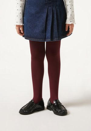 Burgunderfarbene Strumpfhosen, denimfarbene Faltenrock, langärmliges weißes Hemd mit Punkten und schwarze Mokassinschuhe im Fransenstil.