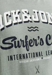 Gaiši zaļš kokvilnas T-krekls ar izteiksmīgu melnu un baltu tekstu, kurā rakstīts 'JACK & JONES' un 'Surfer's Cove', viļņu grafika un zīmola logo.