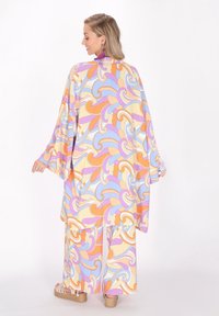 Costum colorat, cu un model, realizat din țesătură fluidă cu valuri portocalii, violet și pastelate. Mâneci largi și o croială relaxată, completat cu pantofi cu talpă înaltă.