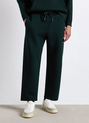 Persona che indossa pantaloni sportivi larghi verde scuro con coulisse in vita e sneakers bianche, in piedi su un pavimento grigio chiaro.
