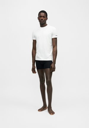 Mann, barfuß stehend, trägt ein weißes T-Shirt mit kurzen Ärmeln und schwarze eng anliegende Shorts vor einem schlichten hellen Hintergrund.