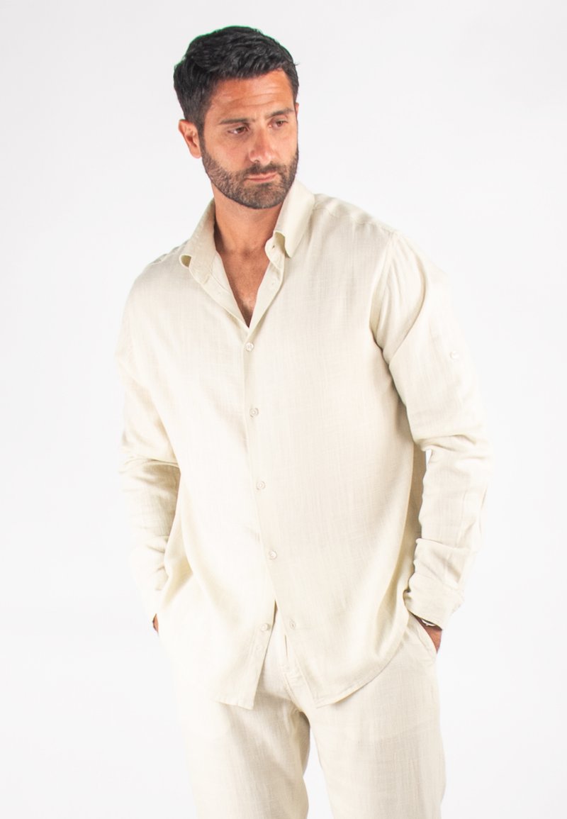 Chemise beige clair à manches longues et boutons, fabriquée en tissu texturé ; poches avant et poignets avec détails en bouton. Portée avec un pantalon assorti.