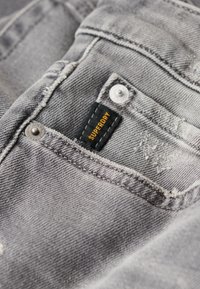 Tela de denim gris con detalles deshilachados, dos botones de metal y una etiqueta negra con texto naranja que dice "SUPERDRY."