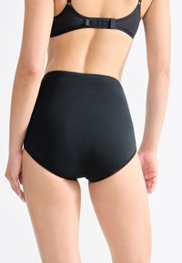 Donna che indossa biancheria intima nera a vita alta e reggiseno nero, vista da dietro su uno sfondo bianco uniforme.