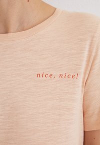 Light peach T-shirt made of soft fabric, featuring a crew neck and the text "nice, nice!" printed in bold red letters on the front.