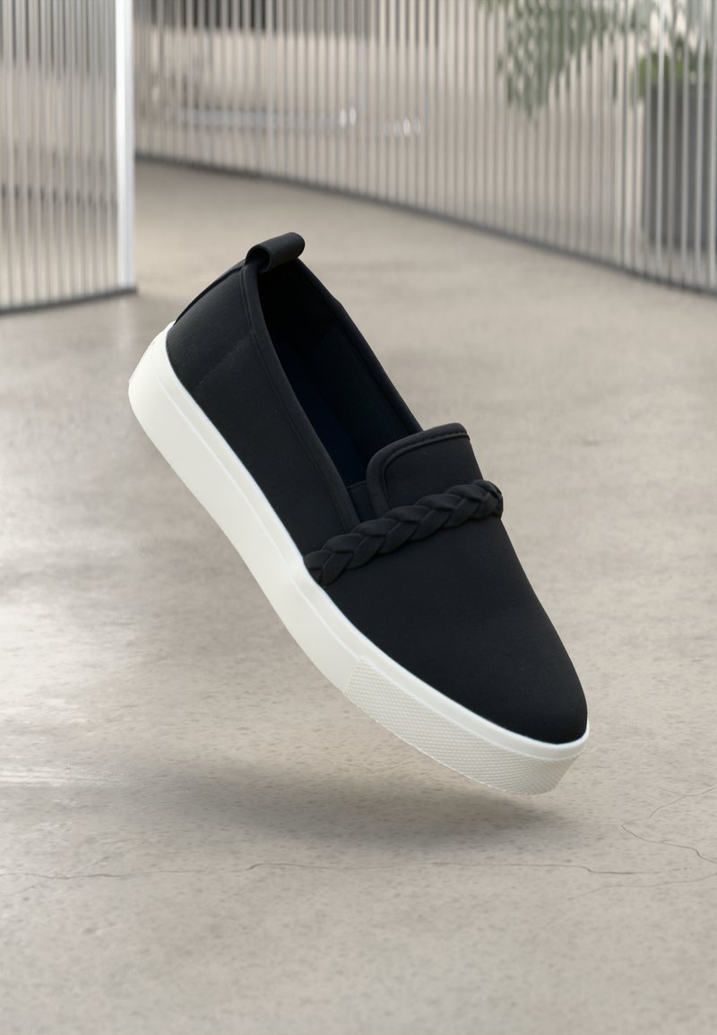Black canvas slip-on shoe featuring a braided accent, white rubber sole, rounded toe, and smooth texture.