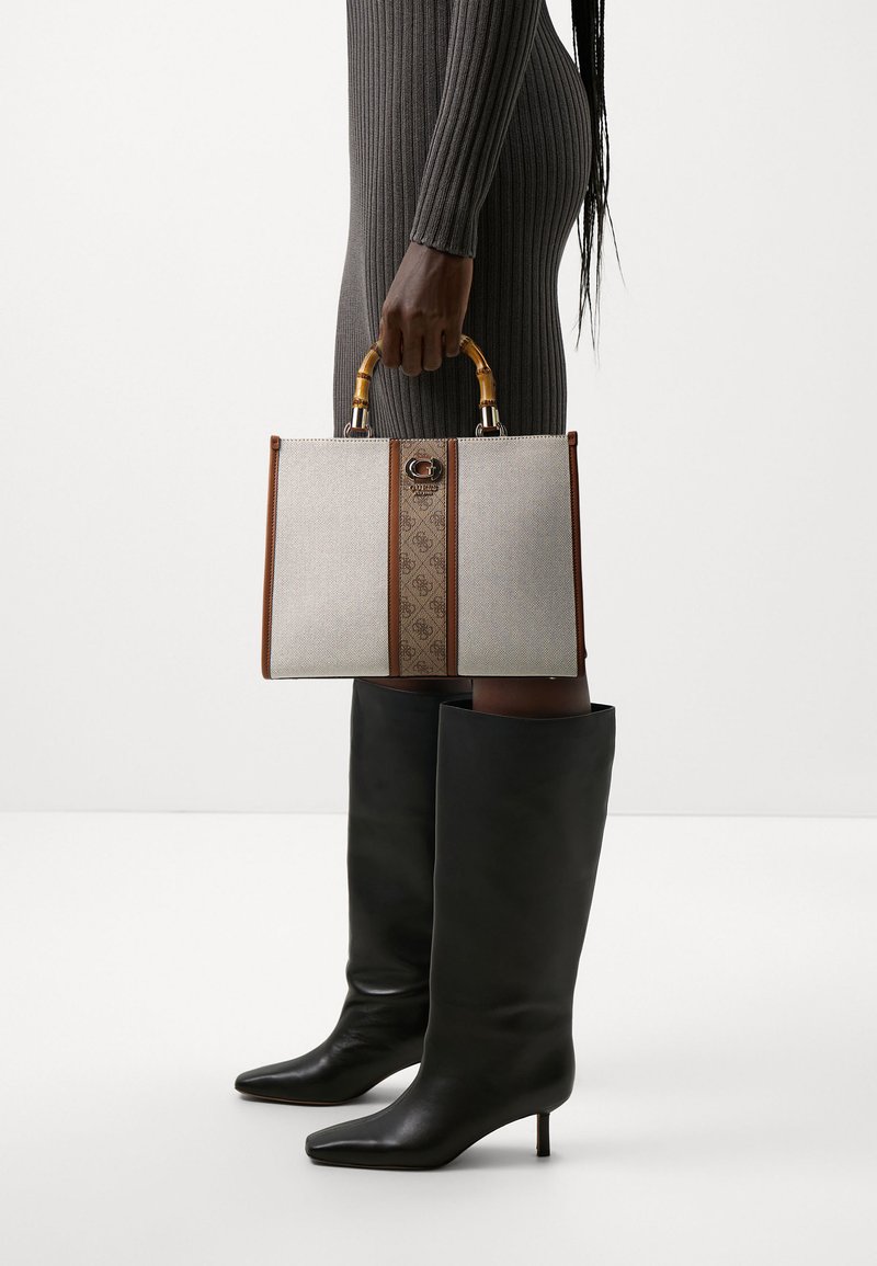 Sac à main beige avec des accents marron, des poignées en bambou et un motif monogramme central. Conception structurée avec une finition texturée.