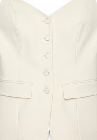 Krämfärgad blazer med v-ringning, sex runda knappar, två framfickor och strukturerade designelement. Slät tygtextur.