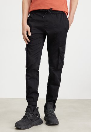 Mężczyzna w czarnych spodniach bojówkach z elastycznymi mankietami i czarnych butach trekkingowych, stojący na jasnej podłodze na białym tle.