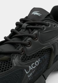 Baskets noires Lacoste avec côtés en mesh, lacets épais, logo crocodile brodé sur la languette et texte de marque sur le côté.