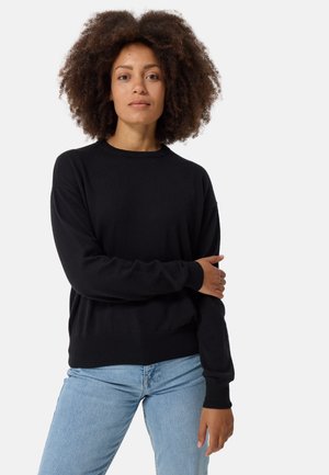 Schwarzer Pullover aus weichem Strick, mit Rundhalsausschnitt, langen Ärmeln sowie gerippten Bündchen und Saum. Kombiniert mit hellblauer Jeans.