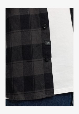 Svart- och grårutigt flanellskjorta med knäppning och logotagg, lager över en vit T-shirt. Texturerat tyg med mjuk yta.