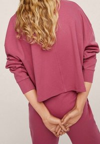 Personne blonde aux cheveux ondulés portant un haut à manches longues rose ample et un pantalon assorti, debout les mains jointes dans le dos.