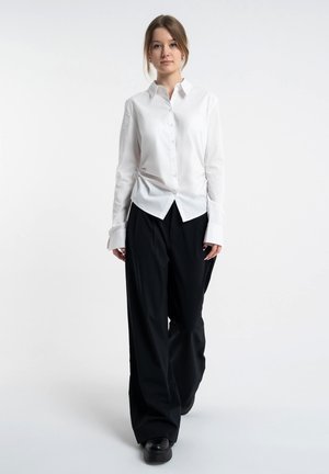 Młoda kobieta stojąca, ubrana w białą koszulę na guziki i luźne czarne spodnie na jednolitym białym tle.