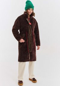 Žena v dlouhém hnědém huňatém kabátu, zelené pletené čepici, krémových kalhotách a hnědých platformových botách, stojící na jednobarevném bílém pozadí.