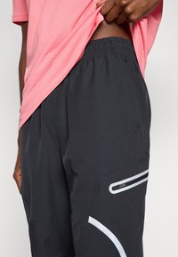 Μαύρο αθλητικό παντελόνι με υφή, ελαστική μέσ η και αντανακλαστικές λευκές λεπτομέρειες κατά μήκος της πλευράς και της τσέπης με φερμουάρ.