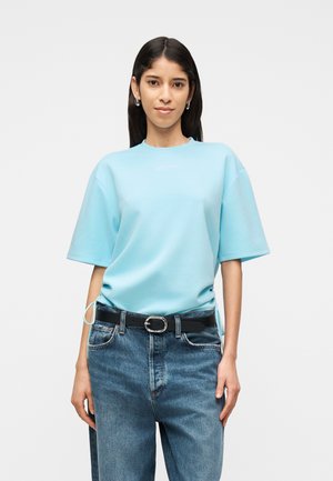 Hosszú sötét hajú nő világoskék, bővített rövid ujjú felsőt visel, amelyet magas derekú kék farmernadrágba tűrt be, fekete övvel.