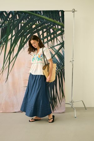 Frau trägt eine weiße floral bestickte Bluse und einen langen blauen Rock, trägt eine geflochtene Strohtasche und steht vor einem großen Palmenblatt-Hintergrund.