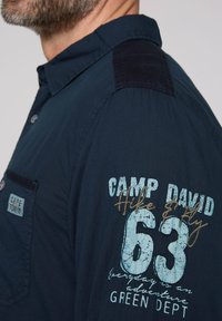 Camicia blu navy con design a bottoni, caratterizzata da una toppa scura sulla spalla, una tasca sul petto con etichetta e testi e grafiche stampate in evidenza.
