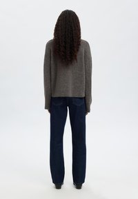 Pilkas megztinis laisvo silueto, derinamas su tamsiai mėlynomis tiesiomis džinsais, matomas iš nugaros priešais paprastą foną.