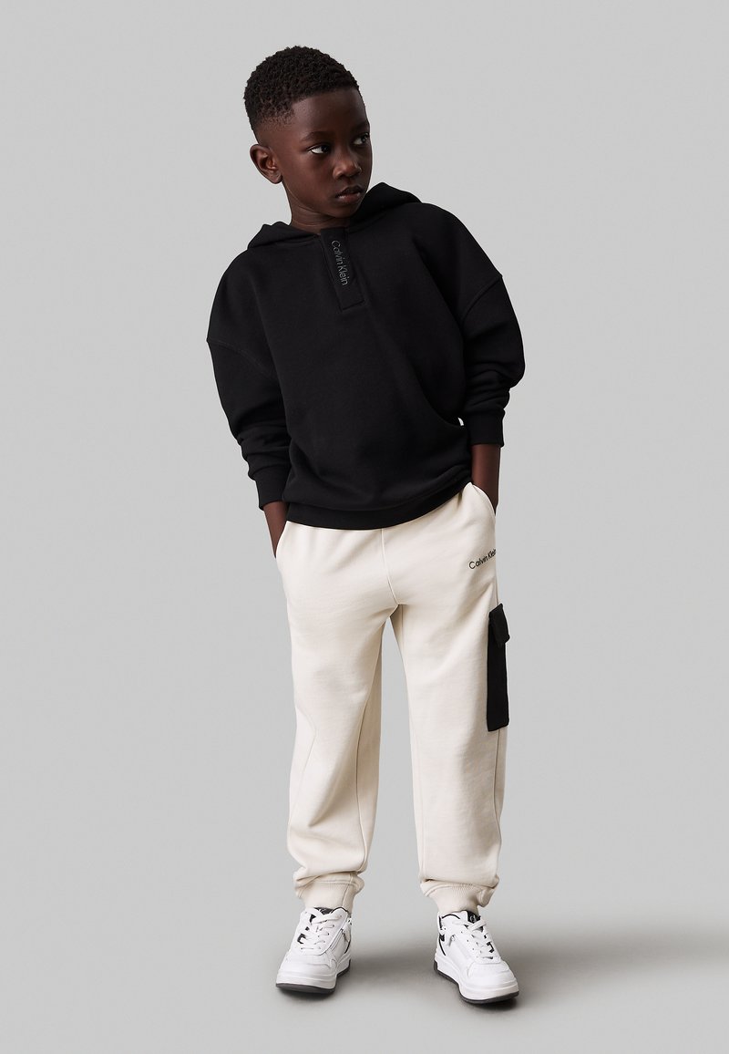 Zwarte hoodie met logo-detail, crèmekleurige joggers met zijzak en witte sneakers. Soepele stof en relaxte pasvorm.