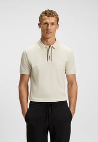 Hellbeige Poloshirt mit umklappbarem Kragen, versehen mit einer schwarz-weißen gestreiften Knopfleiste. Kurze Ärmel und glattes, strukturiertes Gewebe.