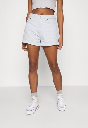 Ung person iført lyseblå opkantsede denimshorts, grå crop top, grå sokker og hvide høje sneakers, stående mod en ensfarvet baggrund.