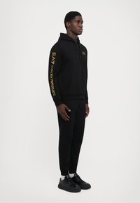 Zwarte hoodie met gouden logo's op de mouwen en op de borst, gecombineerd met zwarte joggers en sneakers. Het materiaal lijkt zacht en heeft een relaxte pasvorm.