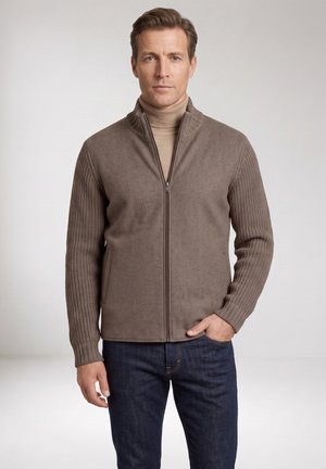 Man wearing a taupe zip-up jacket with knitted sleeves and a beige turtleneck, standing against a plain light background.