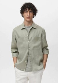 Svijetlozelena košulja sa dugim rukavima, dugmadi, dva prednja džepa i ležernim krojem, preko sive majice, uparena s bijelim hlačama.