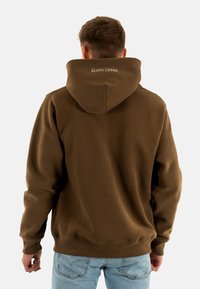 Felpa marrone con un ampio cappuccio, polsini e vita a coste. Presenta un logo ricamato sul retro. Indossata con jeans azzurri chiari.