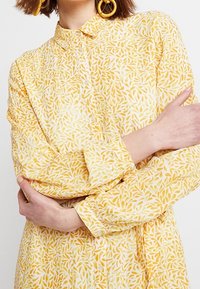 Žlutá košile na knoflíky s květinovým vzorem listů. Má dlouhé rukávy a límec, vyrobená z lehké tkaniny.