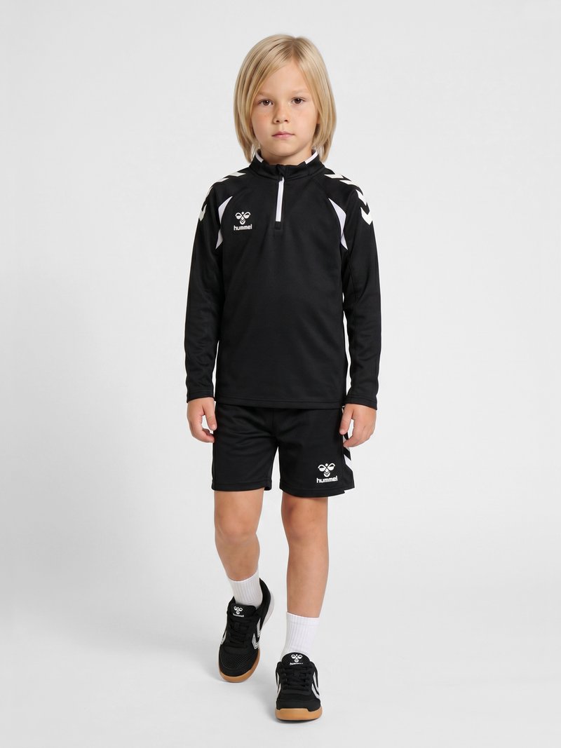 Niño rubio caminando hacia adelante con ropa deportiva negra con detalles blancos, zapatillas negras y calcetines blancos sobre un fondo claro y sencillo.