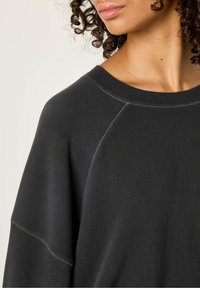 Sweatshirt noir à encolure dégagée, manches raglan et détails de couture visibles. Texture douce avec une coupe décontractée.