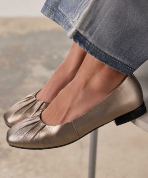 Ballerines métalliques dorées avec un bout rond et des détails plissés. Texture en cuir lisse, petit talon noir, portées avec un jean.