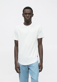 Bílé tričko s krátkým rukávem, kulatým výstřihem a mírně zakřiveným lemem, v kombinaci se světle modrými džínami. Hladká textura látky, bez vzorů.