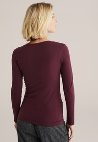 Bordeauxfarbener Langarm-Oberteil mit Rundhalsausschnitt, glatter Textur und tailliertem Design, kombiniert mit dunklen, strukturierten Hosen.