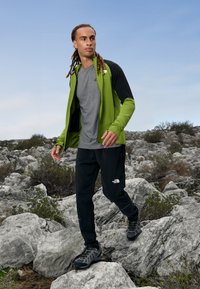 Zielono-czarna kurtka zapinana na zamek, na szarym podkoszulku, w połączeniu z czarnymi spodniami i szarym obuwiem turystycznym, na tle skalistego terenu.