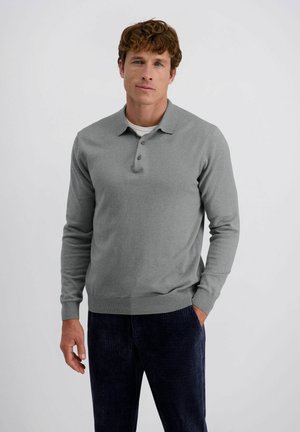 Man in een grijze lange mouw polo en donkerblauwe corduroy broek, staande met één hand in de zak tegen een egale achtergrond.